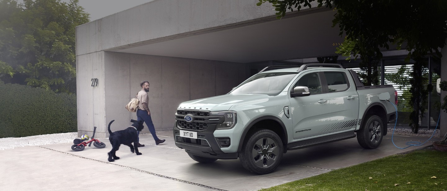 Muž se psem vchází do domu a míjí při tom plug-in hybridní Ford Ranger Stormtrak připojený do domácí nabíječky. Muž se psem vchází do domu a míjí při tom plug-in hybridní Ford Ranger Stormtrak připojený do domácí nabíječky.
